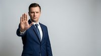 Professional businessman in navy suit making stop gesture with hand raised, serious expression on neutral background for business communication concepts.
