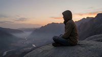 Young man in dark hoodie sitting on mountain cliff edge at sunset, contemplating misty valley landscape below with dramatic peaks and winding river.