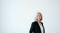 Mature woman with gray hair wearing black blazer and white shirt, smiling at camera against white background. Professional headshot for corporate, business, and executive portraits.