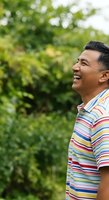 Middle-aged man of Southeast Asian descent wearing colorful striped polo shirt, smiling outdoors with lush green foliage background. Suitable for lifestyle, happiness, and wellness content.