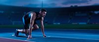 Athletic African American male sprinter in starting position on blue track during twilight training session preparing for competitive race.