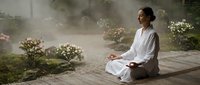 Young Asian woman in white clothing practicing meditation in peaceful zen garden with morning mist and blooming flowers on wooden deck for mindfulness wellness.