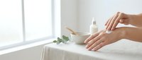 Female hands applying moisturizer cream from jar on white towel in bright spa setting with natural light for skincare routine and beauty treatment.