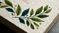 Elegant embroidered botanical leaf branch on textured cream paper, showcasing delicate green and teal foliage stitching for stationery or invitation design.