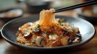 Glass noodles with mushrooms and savory sauce lifted by black chopsticks, steam rising from dark ceramic bowl, garnished with fresh herbs and sesame seeds.
