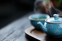 Traditional turquoise ceramic teapot with floral pattern and matching cup on wooden tray, steam rising against dark background creates serene Asian tea ceremony atmosphere.