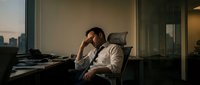 Exhausted businessman sitting at office desk during late evening hours, experiencing work stress and fatigue in corporate environment.