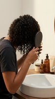 Young woman with curly hair looking in mirror while brushing hair in modern bathroom with skincare products on counter for morning routine.