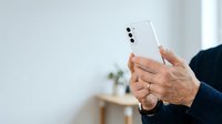 Mature man with medium skin tone holding white smartphone with dual camera setup, wearing dark navy blazer, indoor office environment with blurred background and natural lighting.