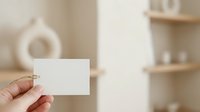 Person holding blank white business card in modern minimalist interior with shelves and home decor, suitable for branding and corporate mockups.