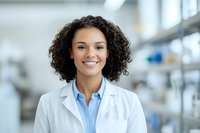Young African American female medical professional with curly hair wearing white lab coat and blue shirt smiling confidently in modern healthcare facility laboratory setting.
