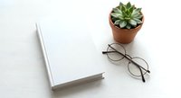 Minimalist workspace with blank notebook, eyeglasses and succulent plant on white desk. Clean office setup for productivity and planning concepts.