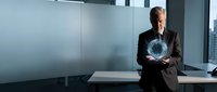 Senior Caucasian businessman in dark suit holding glowing digital sphere in modern office environment with glass walls and contemporary furniture.