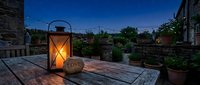 Romantic outdoor patio at dusk with glowing lantern, love stone, rustic wooden table, potted plants, and string lights under a twilight sky.