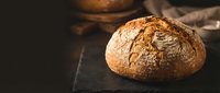 Freshly baked rustic sourdough bread loaf on dark stone board, golden crust, artisan bakery concept with moody dark background.