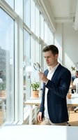 Young Caucasian businessman in navy suit examining documents with magnifying glass in bright modern office with large windows for business analysis.