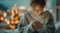 Young blonde woman in cozy knit sweater using smartphone at home with warm bokeh lights creating intimate evening atmosphere for lifestyle content.