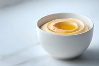 Creamy mayonnaise sauce in white ceramic bowl with olive oil on top, served on marble surface against light background. Fresh homemade condiment.