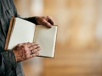 Senior man with weathered hands holding open blank notebook, wearing dark cardigan against warm blurred background. Suitable for education, writing, journaling, and lifestyle concepts.