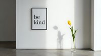 Minimalist interior with a framed be kind motivational poster on a white wall and a yellow tulip in a glass vase casting a soft shadow.
