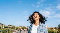 Young woman with dark wavy hair and medium skin tone wearing denim shirt over white top, smiling outdoors with blue sky and urban park background, lifestyle portrait.