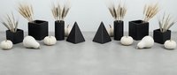 Modern minimalist home decor arrangement with black geometric vases, dried pampas grass, pyramid sculptures and white stones on concrete surface for interior design.