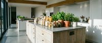 Young woman with light skin tone, wearing casual home attire, arranging potted plants and decorative items on a rustic wooden kitchen island in a modern farmhouse kitchen with white cabinetry and natural light.