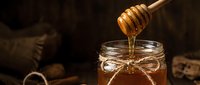 Golden honey dripping from wooden dipper into glass jar with rustic twine bow on dark wooden background with warm lighting for natural food photography.