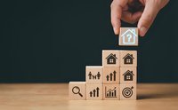 Hand placing wooden cube with house icon on top of stacked blocks showing real estate and business growth symbols on wood table against dark background.