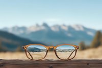 Brown-framed eyeglasses on wooden surface with majestic snow-capped mountain peaks visible through lenses, creating optical illusion against blurred natural landscape background.