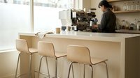 Barista preparing coffee behind counter in modern minimalist cafe with wooden bar seating, bright natural light, contemporary interior design for hospitality and restaurant concepts.