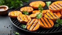 Grilled sweet potato slices with char marks and fresh herbs on black grill pan, garnished with parsley leaves against dark background. Fresh healthy food.