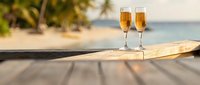 Two champagne flutes filled with golden bubbly on wooden deck overlooking tropical beach with palm trees and turquoise ocean for luxury vacation celebration.
