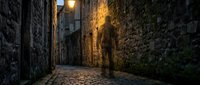 Solitary figure walking through narrow cobblestone alley at dusk, illuminated by warm street lamp glow between ancient stone walls covered in ivy and moss.