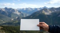 Person holding blank white card with mountain landscape backdrop, suitable for mockups, travel promotions, and outdoor advertising campaigns.