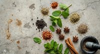Aromatic spices and fresh herbs scattered on textured stone surface with mortar and pestle for culinary cooking and seasoning preparation.