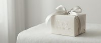 Elegant white gift box with Happy Birthday inscription and satin ribbon bow on soft textured surface, minimal luxury style for celebration.