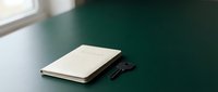 Closed cream notebook and black vintage key on dark teal desk surface near window, minimalist workspace setup for business, education, or creative projects.
