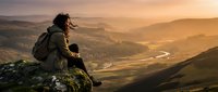 Hiker with backpack sitting on mountain peak overlooking vast valley landscape at golden hour sunset, contemplating scenic wilderness view for travel and adventure concepts.