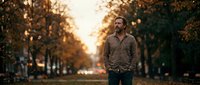 Adult male with light skin tone and beard wearing casual brown jacket standing alone on tree-lined avenue during golden hour sunset, conveying contemplative mood for lifestyle and portrait use.