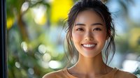 Young Asian woman with long dark hair smiling outdoors in natural sunlight with blurred green foliage background, suitable for lifestyle and portrait use.