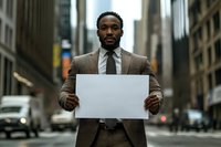 Young African American businessman in brown suit holding blank white sign on urban street with skyscrapers and city traffic in background, serious expression.