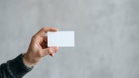 Man with medium skin tone holding blank white business card against neutral gray background for branding and corporate identity mockup purposes.