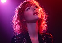 Young woman with red curly hair in sequin jacket illuminated by dramatic red and purple stage lighting, looking up with parted lips in artistic portrait.