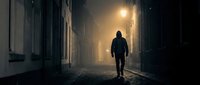 Young adult male figure in dark hoodie walking alone through narrow urban alley at night, illuminated by warm streetlight, creating moody atmospheric scene for thriller, mystery, or crime-related projects.