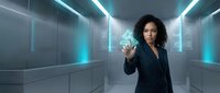 Woman with curly dark hair and medium skin tone in professional business attire pointing at glowing digital interface in modern futuristic corridor with neon lighting and data visualization elements.