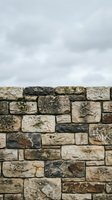 Ancient weathered stone wall with moss and lichen against cloudy sky background for rustic architectural design projects.