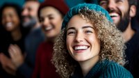 Young woman with curly blonde hair laughing joyfully in crowd, wearing teal sweater. Natural candid moment captures genuine happiness and friendship in dark setting.