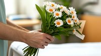 Hand holding fresh bouquet of white daisies with green leaves and decorative ribbon, romantic gesture against blurred interior background, suitable for greeting cards.