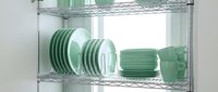 Green ceramic dinnerware set with plates, bowls and cups organized on modern chrome wire shelving rack for kitchen storage and display solutions.